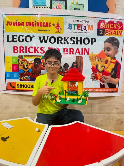 A smiling student points to his LEGO house creation, happy with his completed project.