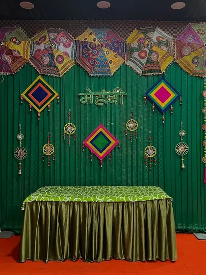 A clean and elegant Mehendi stage with a green backdrop, Rajasthani umbrellas, and colorful hangings, suitable for any venue.
