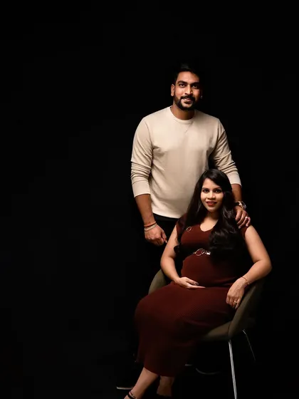 An elegant and classic studio maternity portrait. The simple, dark background and sophisticated lighting create a timeless feel, focusing on the couple's connection and the beautiful silhouette of the baby bump.