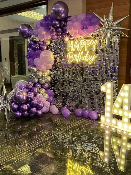 This is a duplicate of ID 64, 129, 145, 159, 196. A glamorous 14th birthday setup with a purple shimmer wall. This decoration features a stunning organic balloon garland in shades of purple and silver, a 'Happy Birthday' neon sign, and large marquee numbers.