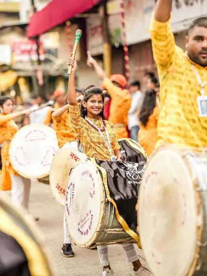 A female dhol player raises her stick high, her smile showing the pride and energy of the performance.