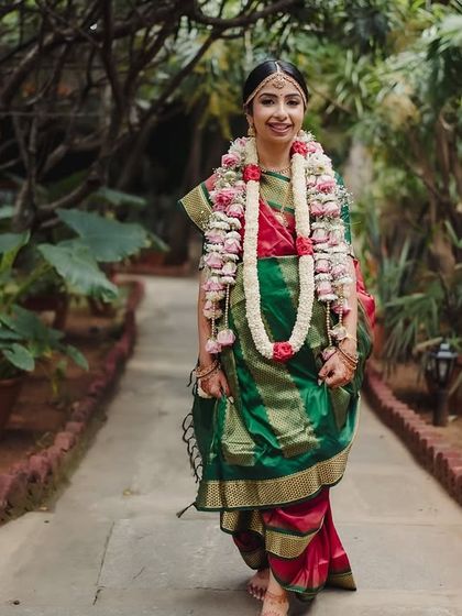 A bride walks down a garden path, her green and red saree vibrant against the lush foliage.