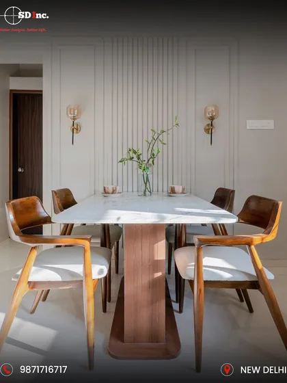 A contemporary dining setup featuring a white marble-top table with a unique wooden base. The chairs, with their elegant wooden frames and light upholstery, complement the fluted wall paneling and classic sconce lights.