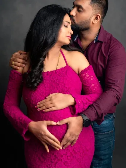 A tender moment with the partner's hands forming a heart on the baby bump. The mom-to-be is wearing a vibrant magenta lace gown.