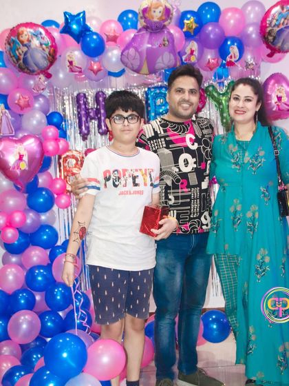 A family poses for a photo against a vibrant pink and blue balloon wall at a birthday celebration.