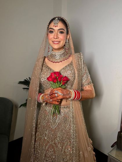 A full shot of the new-age bride in her gold ensemble, holding a bouquet of red roses. The makeup is soft, glowing, and absolutely perfect.