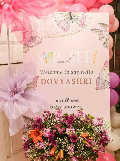 A 'Sip and See' welcome sign for a butterfly-themed baby shower. The sign is adorned with sheer fabric flowers and surrounded by lush floral arrangements.