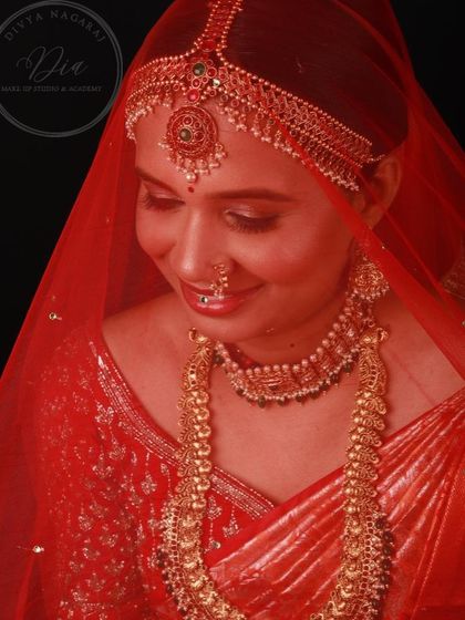 A beautiful shot of a model under a red veil, showcasing a signature South Indian bridal look. The lighting creates a soft, ethereal glow, highlighting the intricate jewellery and the radiant makeup.