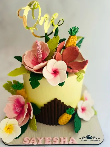 Celebrating a first birthday with a vibrant tropical-themed cake. This detailed creation features handcrafted sugar hibiscus flowers, plumeria, and miniature pineapples, all surrounding a custom gold acrylic "One" topper.