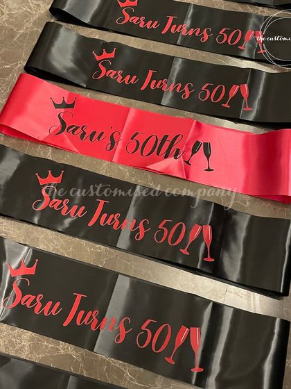 Make the birthday star feel extra special with our custom sashes. These black and red satin sashes are personalized for "Saru's 50th," complete with crowns and champagne glass motifs for a celebratory touch.