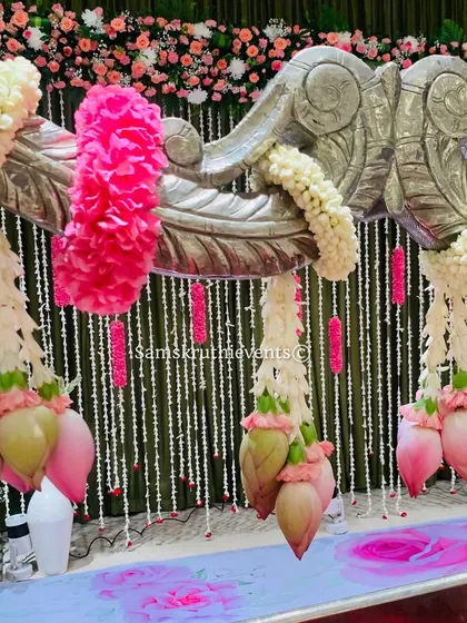 A close-up of a silver cradle decorated with hanging lotus buds and pink floral garlands. The backdrop of hanging jasmine strings adds a delicate and fragrant touch to the setup.