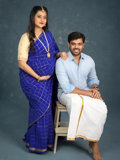 A classic couple portrait from a South Indian traditional maternity shoot. The mom-to-be is radiant in her blue saree, and her partner is dressed in a simple shirt and veshti.
