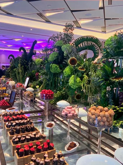 This is my vision of a garden of sweetness. I designed this expansive dessert display for a corporate gala, using lush foliage and dramatic floral structures to frame an abundant selection of handcrafted chocolates, tarts, and confections, all illuminated by soft purple lighting.