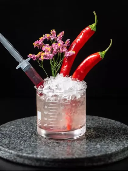 A spicy cocktail served in a beaker glass, garnished with fresh chilies and flowers, with a syringe of red syrup on the side. This presentation is a perfect blend of our 'Bar Doctor' theme and our love for bold flavors.