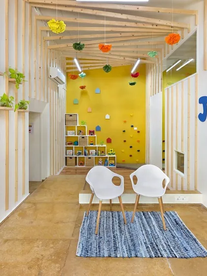The multipurpose entrance lobby of JOY Preschool, which serves as both a reception area and a space for reading sessions. The design features natural wood slats, custom child-safe furniture, and a small climbing wall against a vibrant yellow backdrop to create an engaging and playful atmosphere.