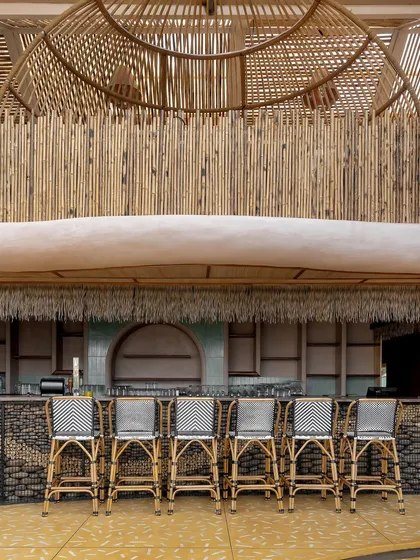 The bar at Dubki features a backdrop of vertically arranged bamboo poles, creating a textured and natural surface. The bar stools are made of cane, continuing the theme of using sustainable and locally relevant materials.