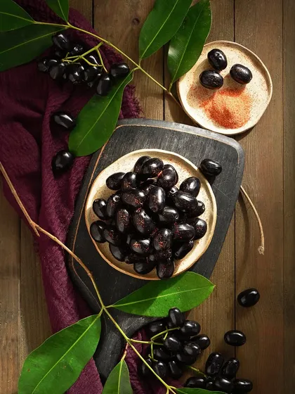 This top-down food flat lay highlights the deep, glossy purple of the Jambhul. The composition plays with different textures, from the smooth berries to the wooden board and fabric.