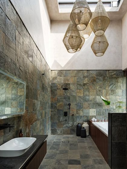 The bathroom is a luxurious, sky-lit space with floor-to-ceiling slate tiles and a cluster of woven pendant lights hanging above the bathtub.