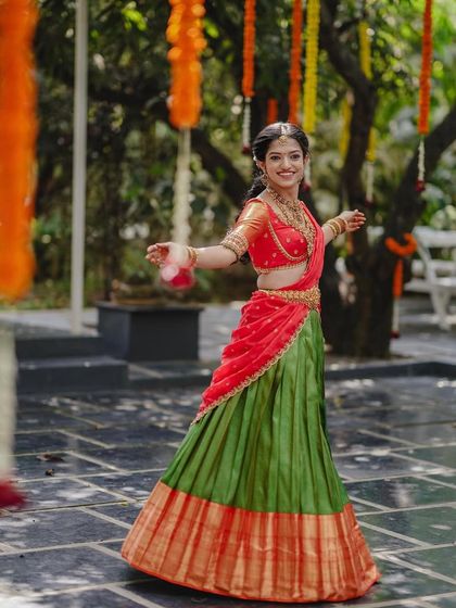 A bride twirls in her colorful half-saree during her engagement ceremony. The courtyard, decorated with hanging marigolds, adds to the festive atmosphere.
