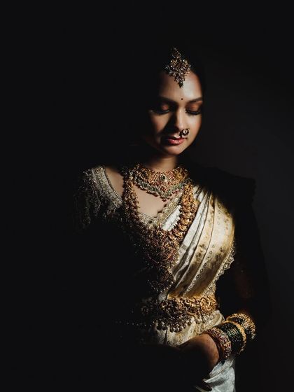 A low-key portrait of a bride, with shadows creating a sense of mystery and elegance. The focus is on the texture of her saree and the sparkle of her jewelry.