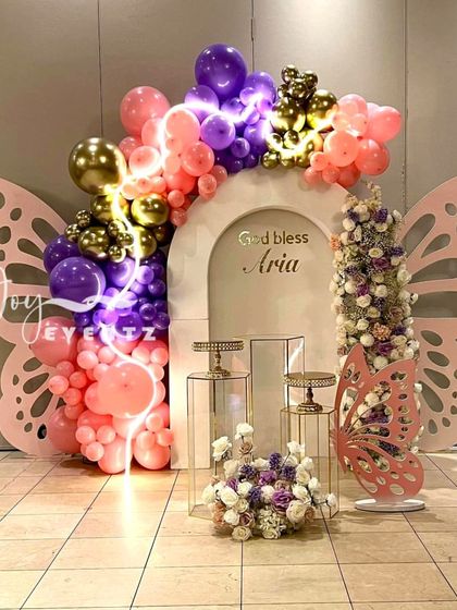 A vibrant butterfly-themed decoration for a First Holy Communion. This design features large pink butterfly wings, a colorful balloon arch with neon light accents, and clear plinths for display.