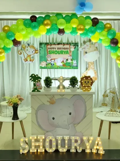A fun and friendly jungle safari theme for a first birthday. This decoration includes a vibrant green and gold balloon arch, cute animal cutouts like a giraffe and lion, and a custom-designed dessert table featuring a baby elephant.