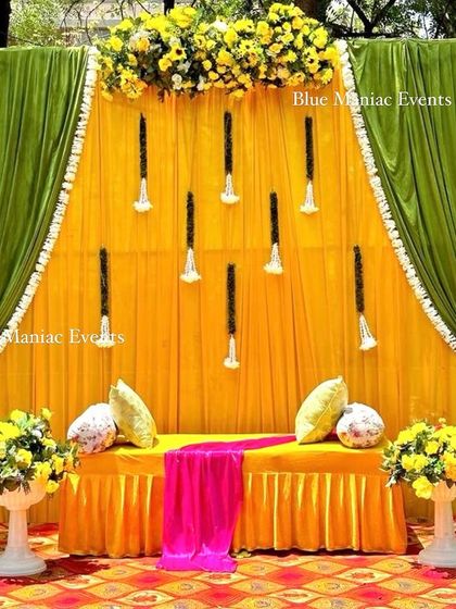 A classic Mehendi and Haldi stage with green and yellow drapes, floral arrangements, and traditional white urns. The patterned carpet adds another layer of texture.