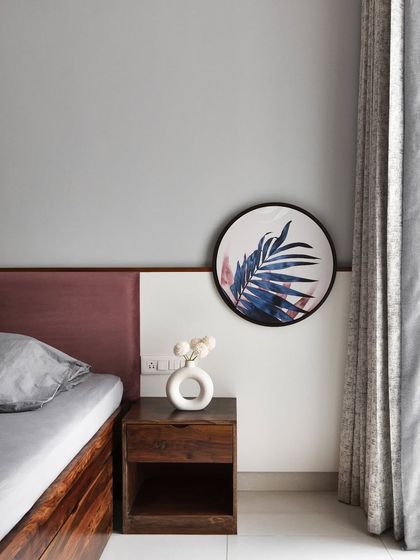 This bedroom uses a soft and soothing pastel color palette. The wall behind the bed features a simple two-tone design with a mauve-colored headboard, complemented by a minimalist wooden side table and a modern art piece.