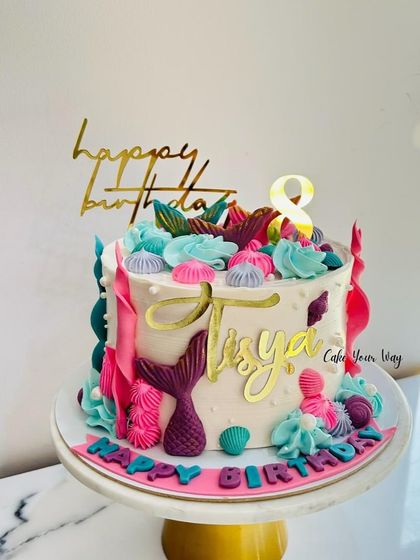 A pretty mermaid-themed cake with vibrant pink and teal frosting, decorated with shells and mermaid tails.
