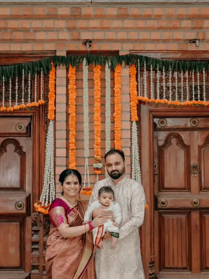 A classic family portrait of the new parents with their baby, taken at their Bangalore home. The traditional wooden doors and marigold decorations create a perfect backdrop for a timeless family photograph.