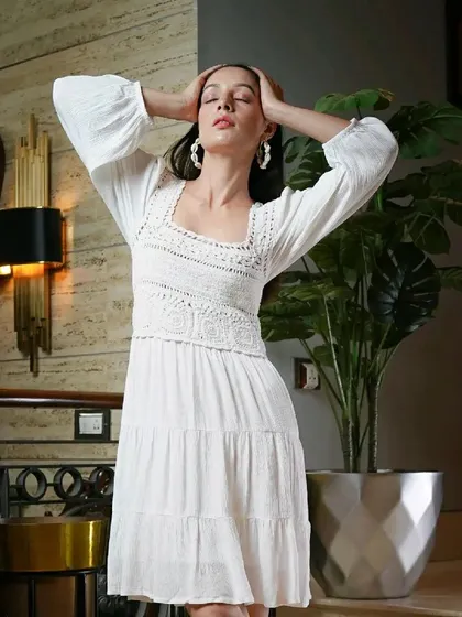 A model strikes a relaxed yet chic pose in a white crochet-detailed dress. The indoor setting with modern decor provides a sophisticated backdrop for this contemporary fashion look.