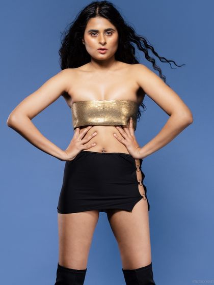 A direct, full-body shot of a model in a gold top and black skirt against a blue background.