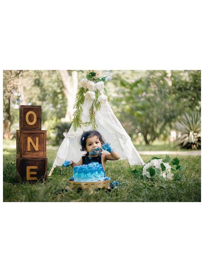 A wide shot of the outdoor cake smash setup, featuring a baby girl, her blue cake, a white teepee, and "ONE" blocks. This shows the complete, beautifully styled scene we create.