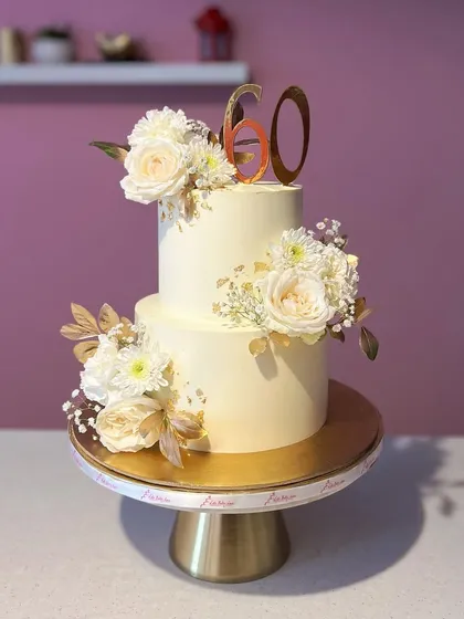 Simple, elegant, and timeless. This two-tiered 60th birthday cake in white and gold is adorned with fresh white roses and a touch of gold leaf, proving that less is often more.