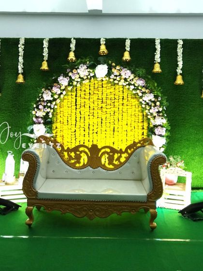 A beautiful stage setup for a Langa Voni function featuring a green turf backdrop and a floral arch. The design includes a yellow marigold curtain and hanging temple bells for a divine touch.