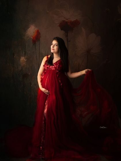 This portrait has a dark, moody elegance. The mom-to-be stands in a flowing red gown against a painted floral backdrop, creating a timeless, artistic feel.
