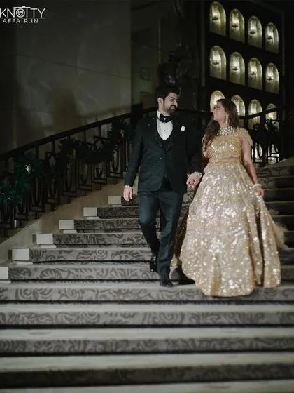 A grand exit for a grand reception. The couple descends the elegant staircase, hand in hand, dressed in their glamorous reception outfits. This shot captures the sophistication and celebratory feel of the evening.
