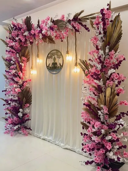 For a modern Jain ceremony, I designed this tranquil backdrop. It features a cascade of pink and white cherry blossoms over a clean white curtain, with warm Edison bulbs and a central laser-cut emblem to create a peaceful and spiritual ambiance.