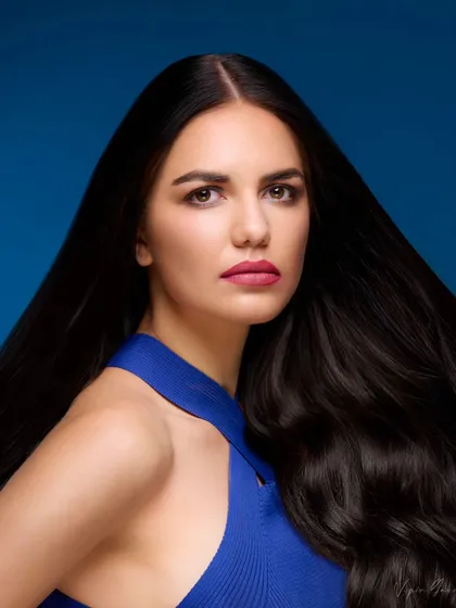 A hair campaign shot focusing on volume and shine. The model's long, dark hair is captured in motion against a solid blue background, emphasizing its health and beauty.