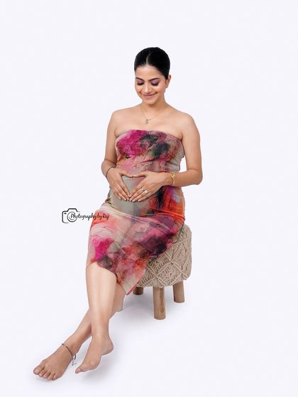 A happy and relaxed studio portrait of a mother-to-be in a colorful abstract dress, seated on a simple white block.