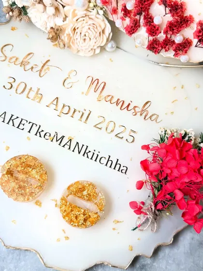 A close-up of a beige-themed ring platter with bold red flower accents. The couple's names and wedding hashtag are featured in gold, and the ring holders are filled with gold flakes.