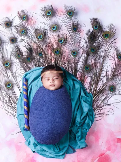 A variation of the Little Krishna theme, with the baby wrapped in blue and surrounded by a fan of peacock feathers on a soft pink background.