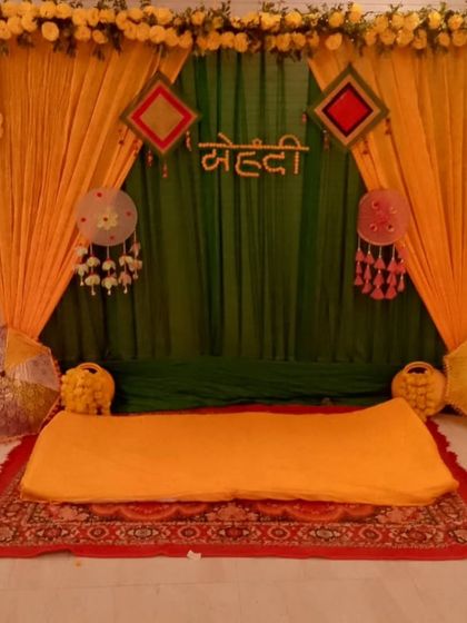 A simple low-seating or gadda setup for a home Mehandi ceremony. The backdrop of green and yellow drapes with a "Mehandi" sign and traditional hangings creates a cozy corner.