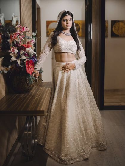A full-length portrait of the bride, Prachi, in her elegant white lehenga. Her poised stance and the classic interior setting create a sophisticated image.