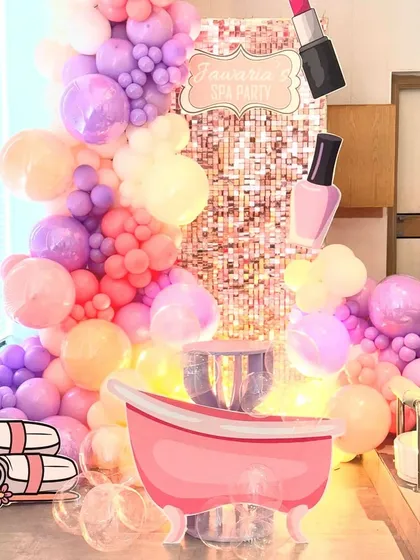 A fun and fabulous spa party theme, perfect for a pamper-filled birthday celebration. This setup includes a shimmering rose gold sequin backdrop, a massive pastel balloon garland, and adorable props like a pink bathtub, lipstick, and nail polish cutouts.