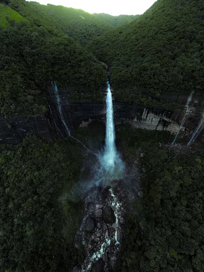 A single, powerful stream of water plunges into a rocky gorge in Meghalaya. This aerial shot highlights the simple, raw beauty of the region's waterfalls.