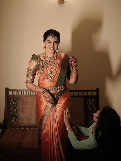 The smiles say it all. A candid moment of joy with a bride after her draping is complete, showcasing a perfect drape on her orange silk saree.