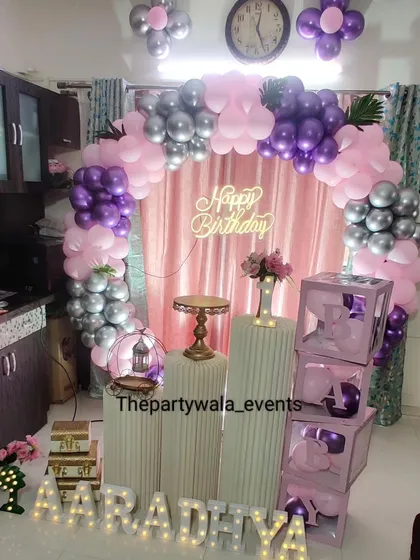 A cozy at-home birthday setup with a pink, purple, and silver balloon ring. The "BABY" blocks and light-up name make the space feel festive.