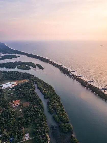 A wide aerial view of the Tulunadu coastline, showing the unique geography where a river runs parallel to the sea, separated by a narrow strip of land.