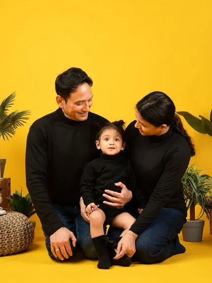 A modern family portrait with a pop of color. The family's coordinated black outfits look striking against the vibrant yellow background and green plants.
